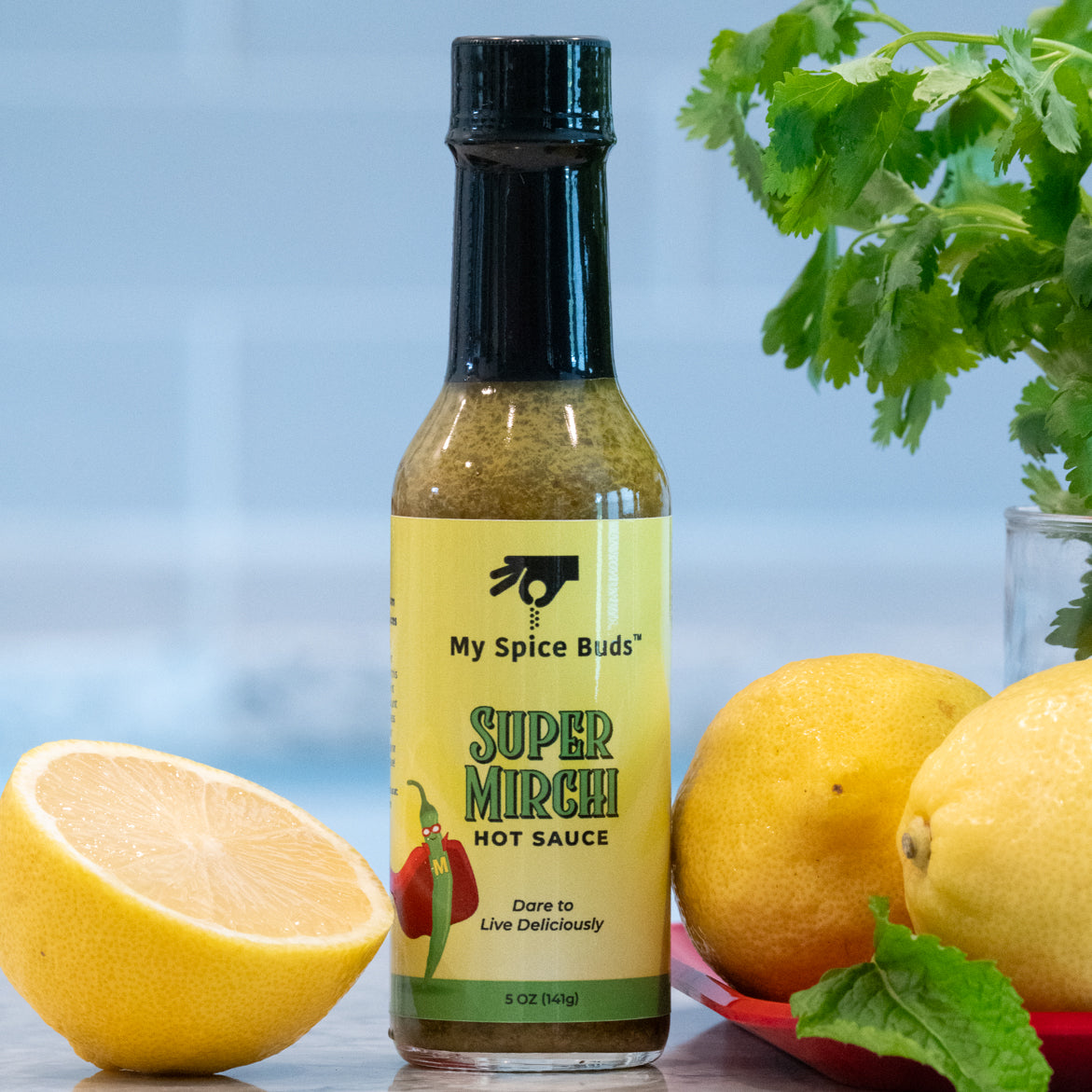 Bottle of My Spice Buds Super Mirchi Hot Sauce with lemons and herbs on a blurred background
