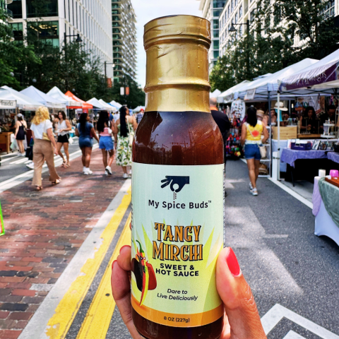 Bottle of 'My Spice Buds Tancy Mirch Sweet & Hot Sauce' held in front of a bustling street market.