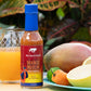 Mango Mirchi hot sauce bottle with a glass of orange juice and mangoes on a table.