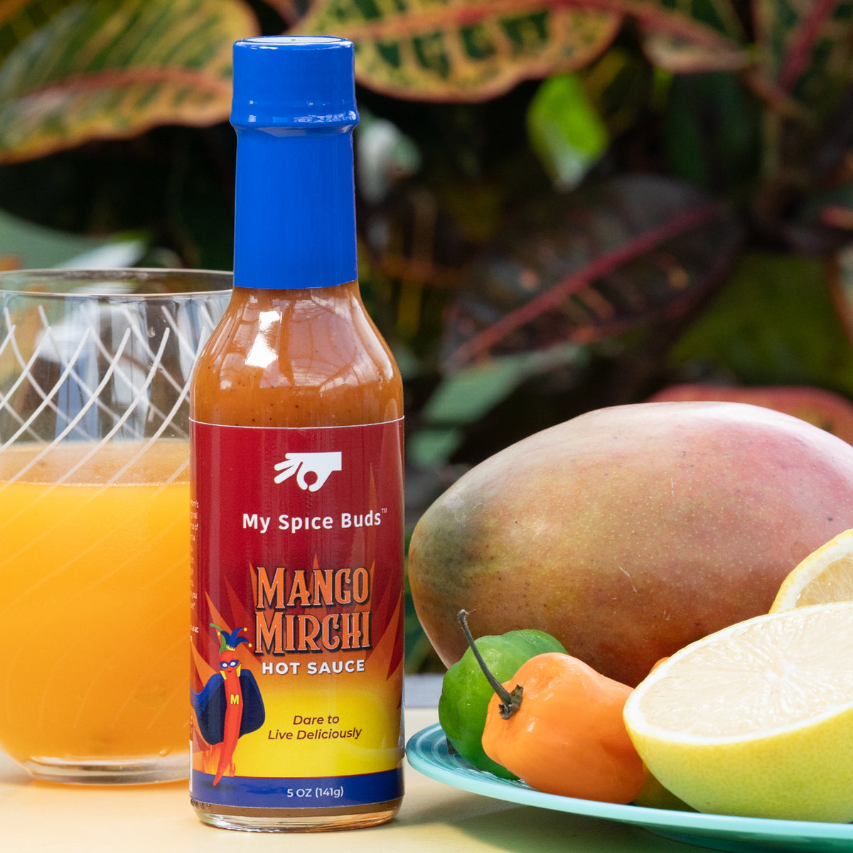 Mango Mirchi hot sauce bottle with a glass of orange juice and mangoes on a table.
