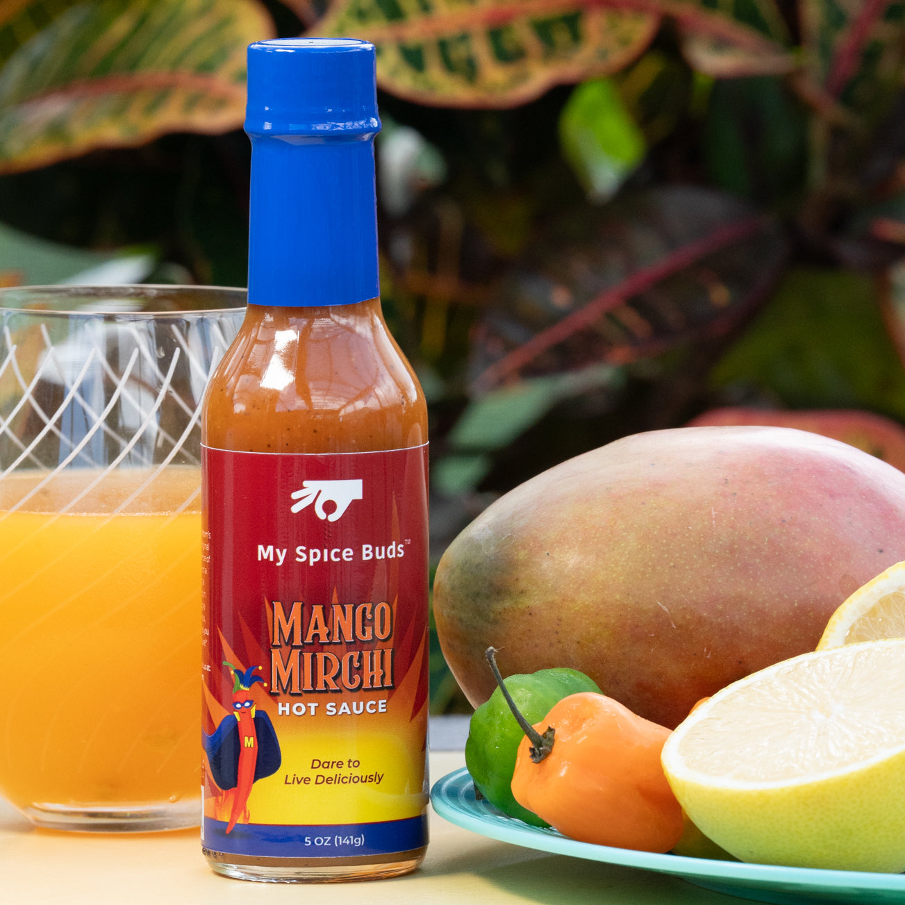 Mango Mirchi hot sauce bottle with a glass of orange juice and mangoes on a table.