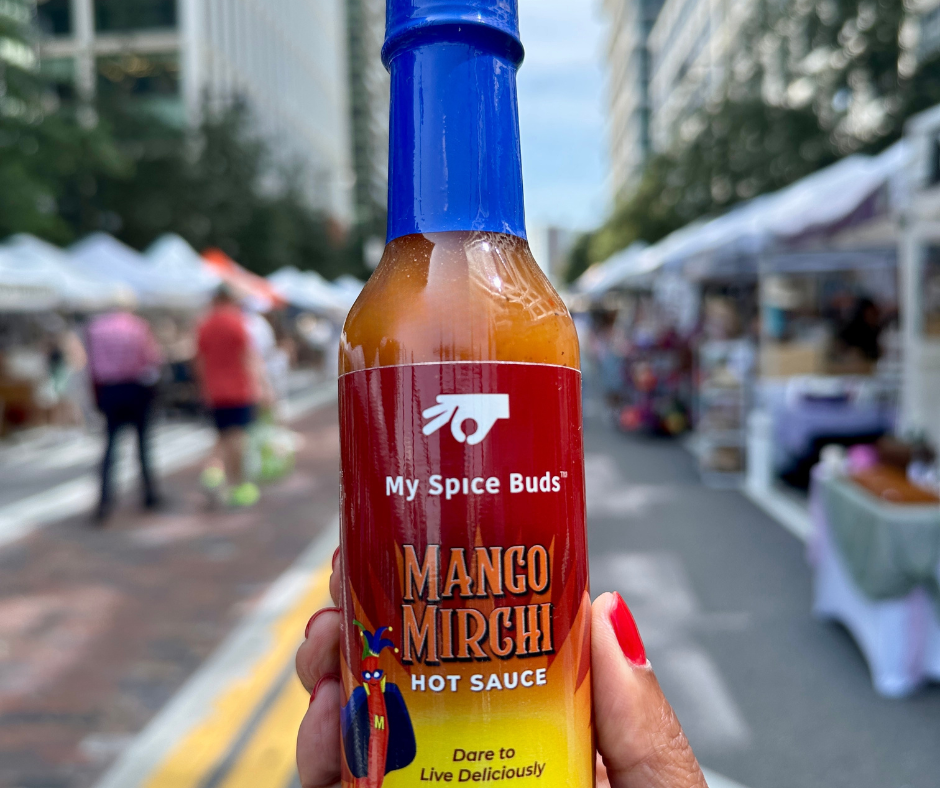 Hot sauce bottle labeled 'My Spice Buds Mango Mirchi Hot Sauce' held in front of a blurred outdoor market scene.