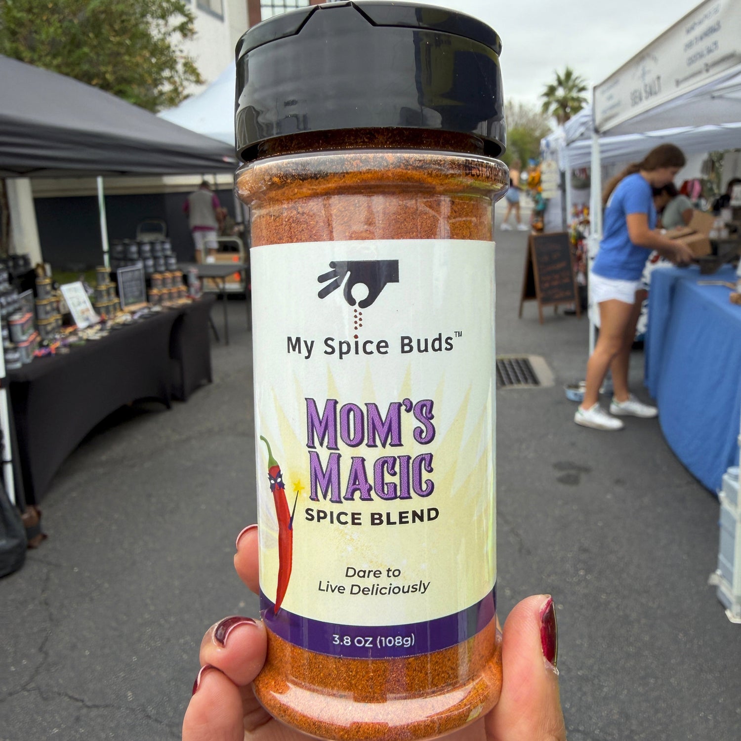 Spice blend jar labeled 'Mom's Magic' held in front of a market stall.