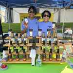 Two women behind a table displaying various bottles and a box of food products under a blue tent. 8