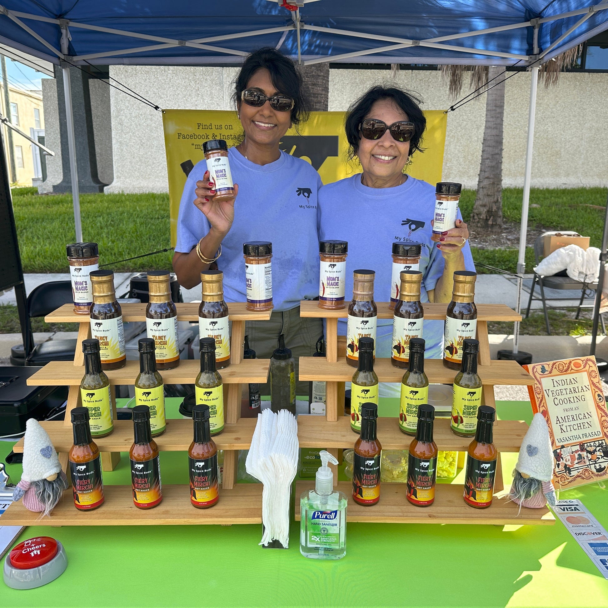 Two women behind a table displaying various bottles and a box of food products under a blue tent.