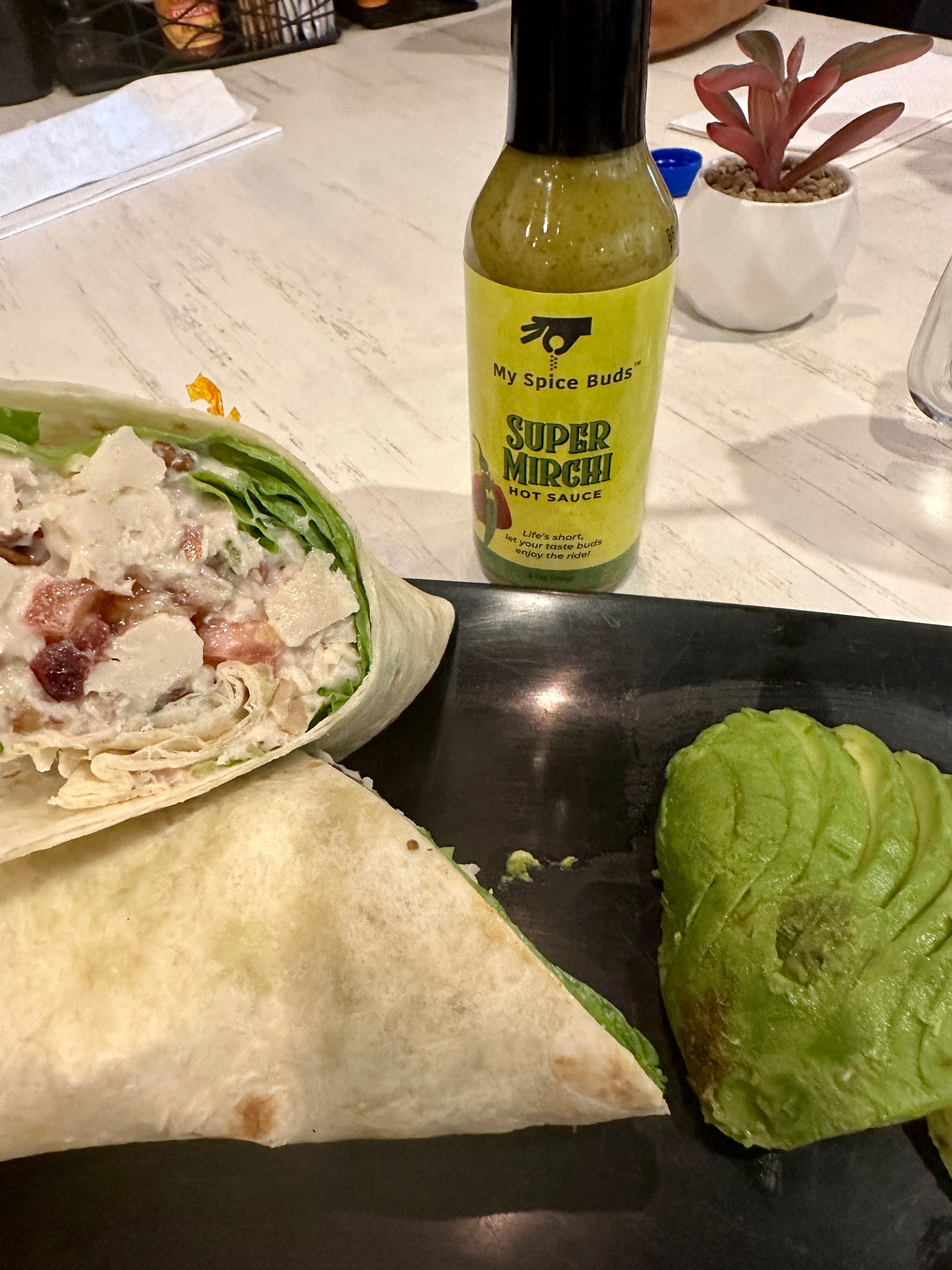 Bottle of "My Spice Buds" Super Much Hot Sauce' on a table with wraps and avocado.